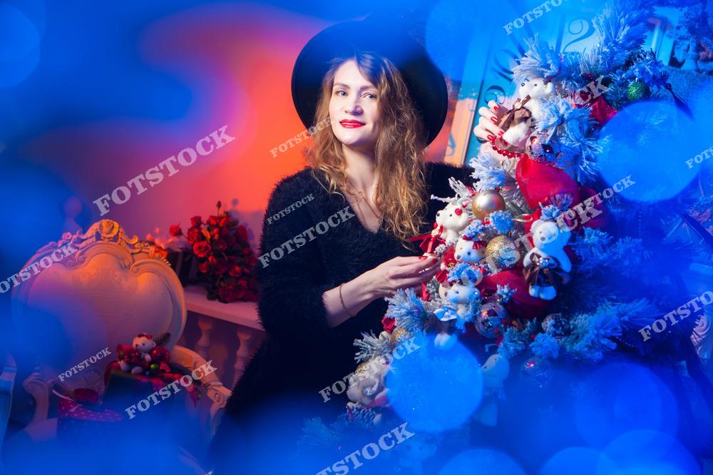 На фотографии изображена женщина, которая украшает рождественскую елку игрушками и гирляндами. Она выглядит счастливой и сосредоточенной на процессе, а окружающая обстановка наполнена праздничным декором. Освещение создает ощущение волшебства и теплоты зимнего праздника.